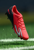 Helle, rote Fußballschuh mit schwarzer Ferse und silbernem Logo, der über Gras auf einem markierten Sportfeld fliegt.
