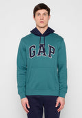Junger Mann, der einen türkisfarbenen GAP-Hoodie mit dunkelblauer Kapuze und dunkelblauen Hosen trägt, steht vor einem einfachen hellgrauen Hintergrund.