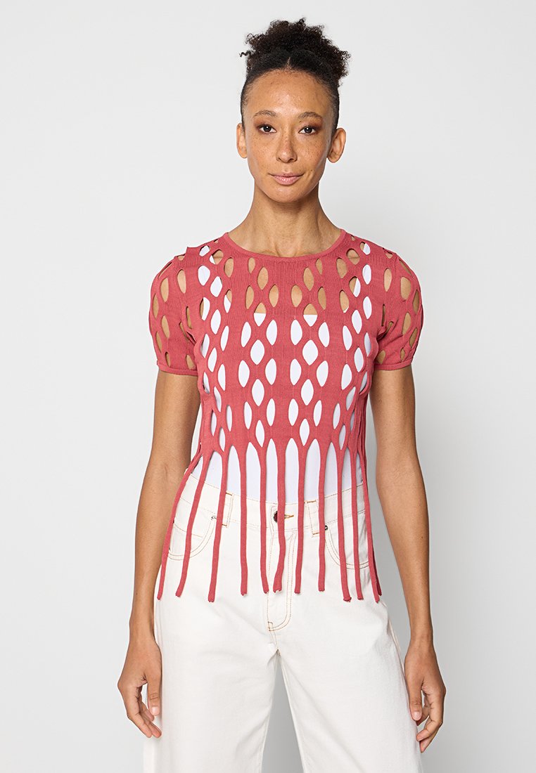 Rotes Strickoberteil mit offenen ovalen Ausschnitten, kurzen puffärmel und einem geschichteten Design mit weißem Unterlayer. Kombiniert mit hellen Hosen.