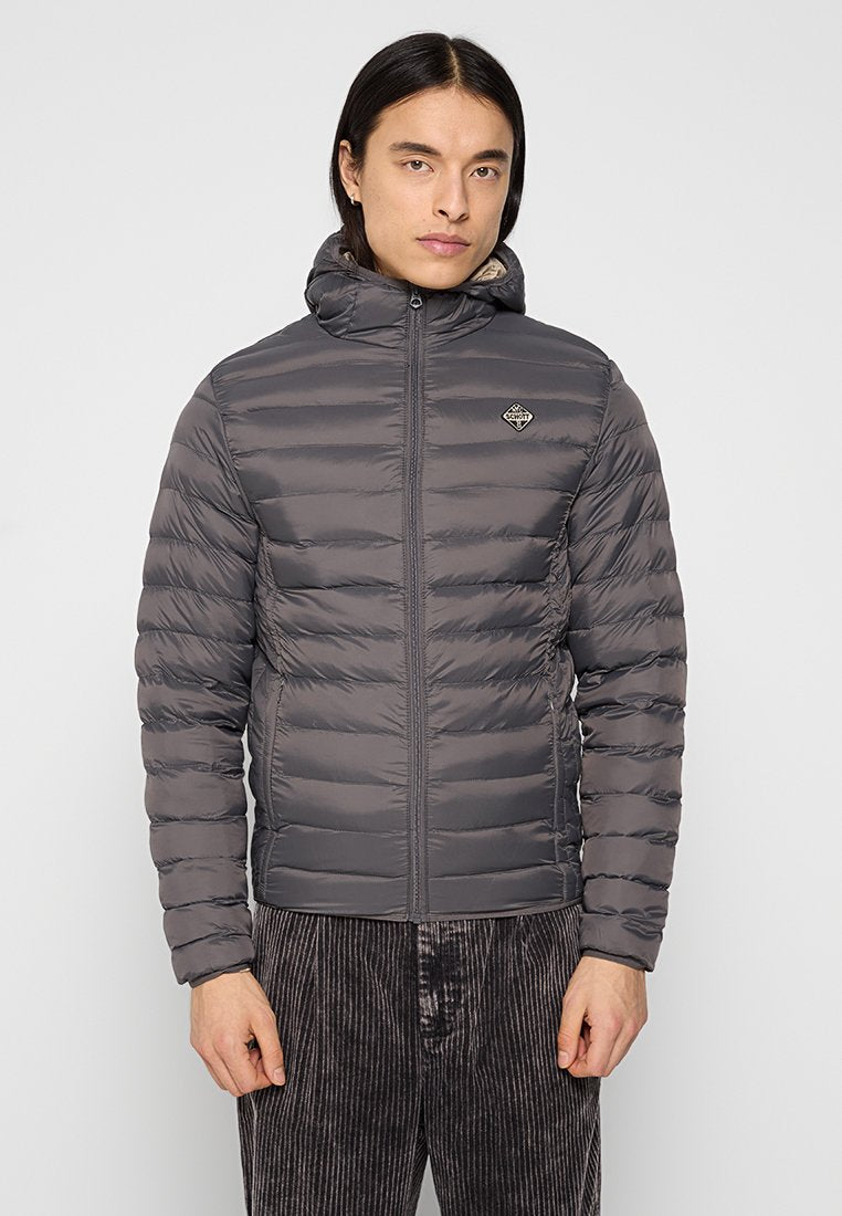 Mann in grauer, wattierter Jacke mit Reißverschluss und Kapuze sowie kleinem Logo auf der Brust, steht vor einem schlichten, hellen Hintergrund.