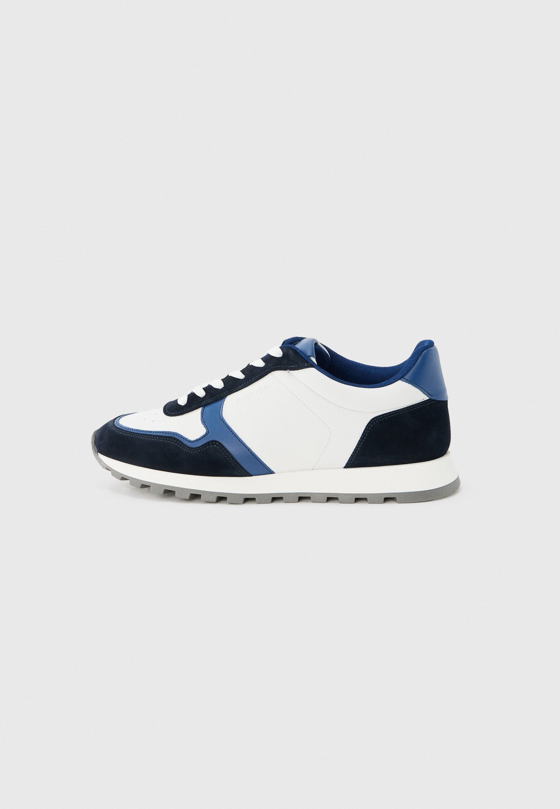 Flacher Sneaker mit weißen, dunkelblauen und grauen Einsätzen, weißen Schnürsenkeln und einer gerillten grauen Sohle auf hellgrauem Hintergrund.