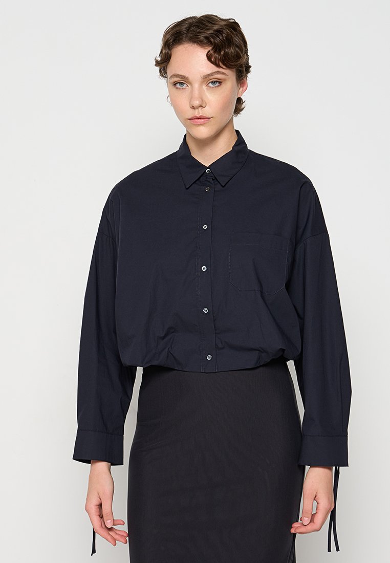 Dunkelblaues Button-Up-Shirt mit spitzem Kragen, langen, lockeren Ärmeln, einer Brusttasche und gerafften Details an den Manschetten.