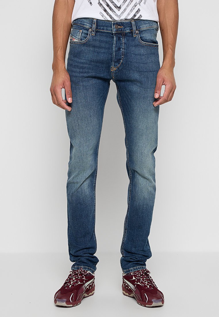 Blaue Denim-Jeans mit schmaler Passform, leichter Ausbleichung an den Beinen, fünf Taschen und kontrastierenden Nähten, kombiniert mit burgunderroten Sportschuhen.