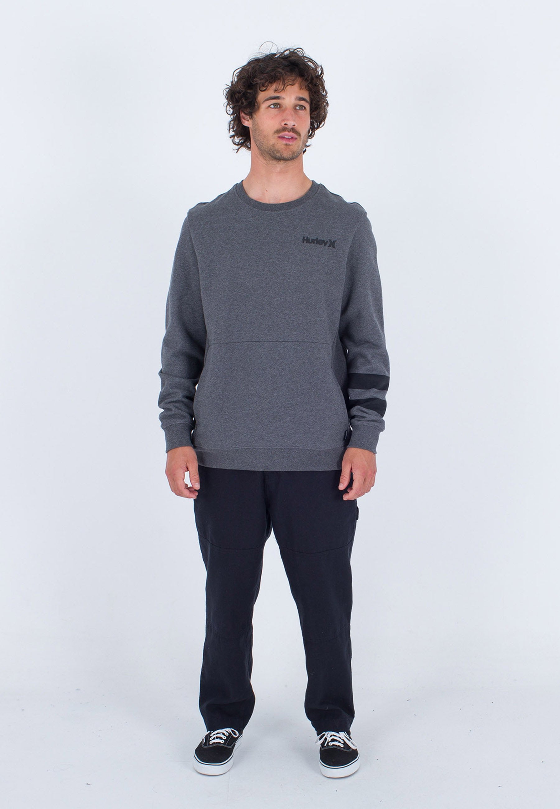 Grauer Sweatshirt mit rundem Halsausschnitt und Schulterpaneelen. Schwarzes Logo auf der Brust. Kombiniert mit dunklen Hosen und schwarzen Sneakers.