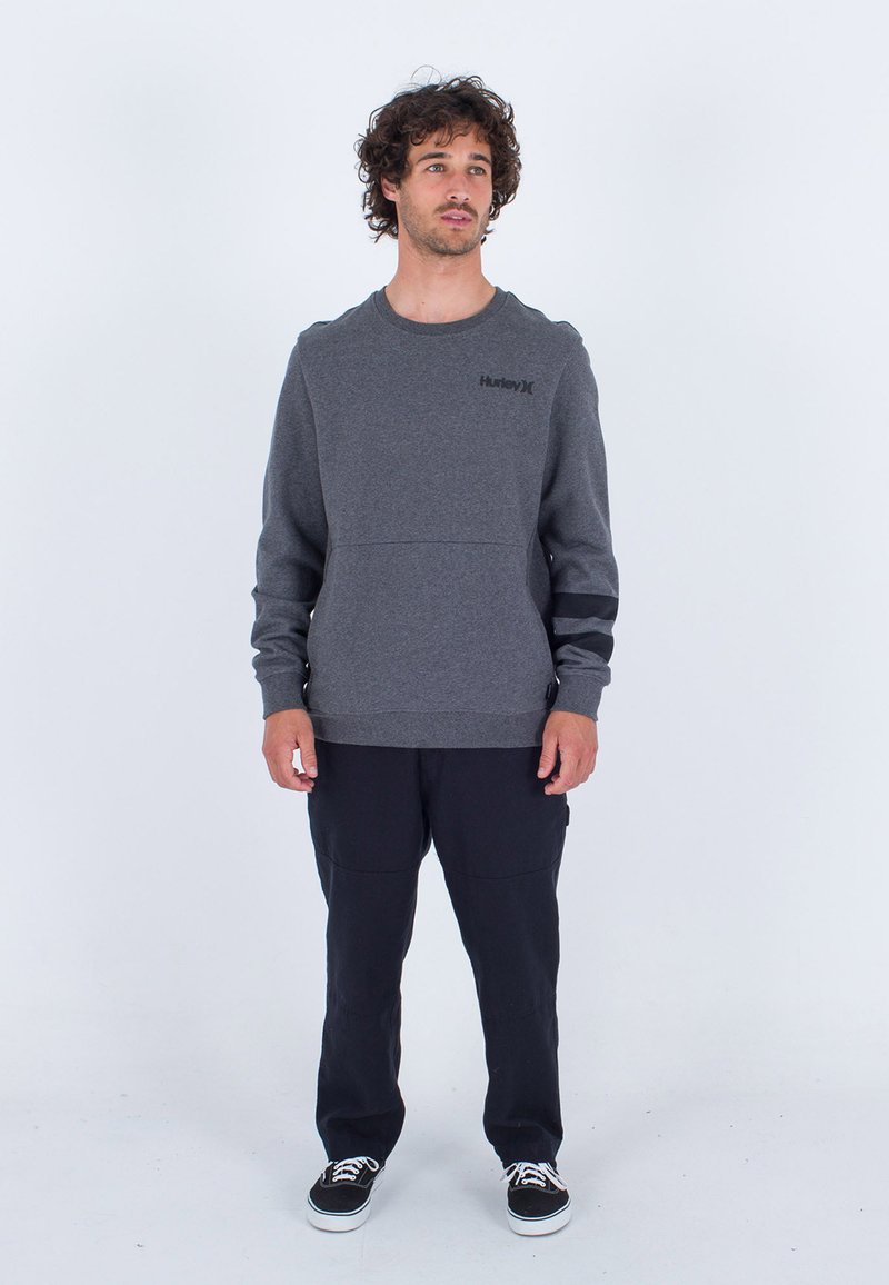 Grauer Sweatshirt mit rundem Halsausschnitt und Schulterpaneelen. Schwarzes Logo auf der Brust. Kombiniert mit dunklen Hosen und schwarzen Sneakers.