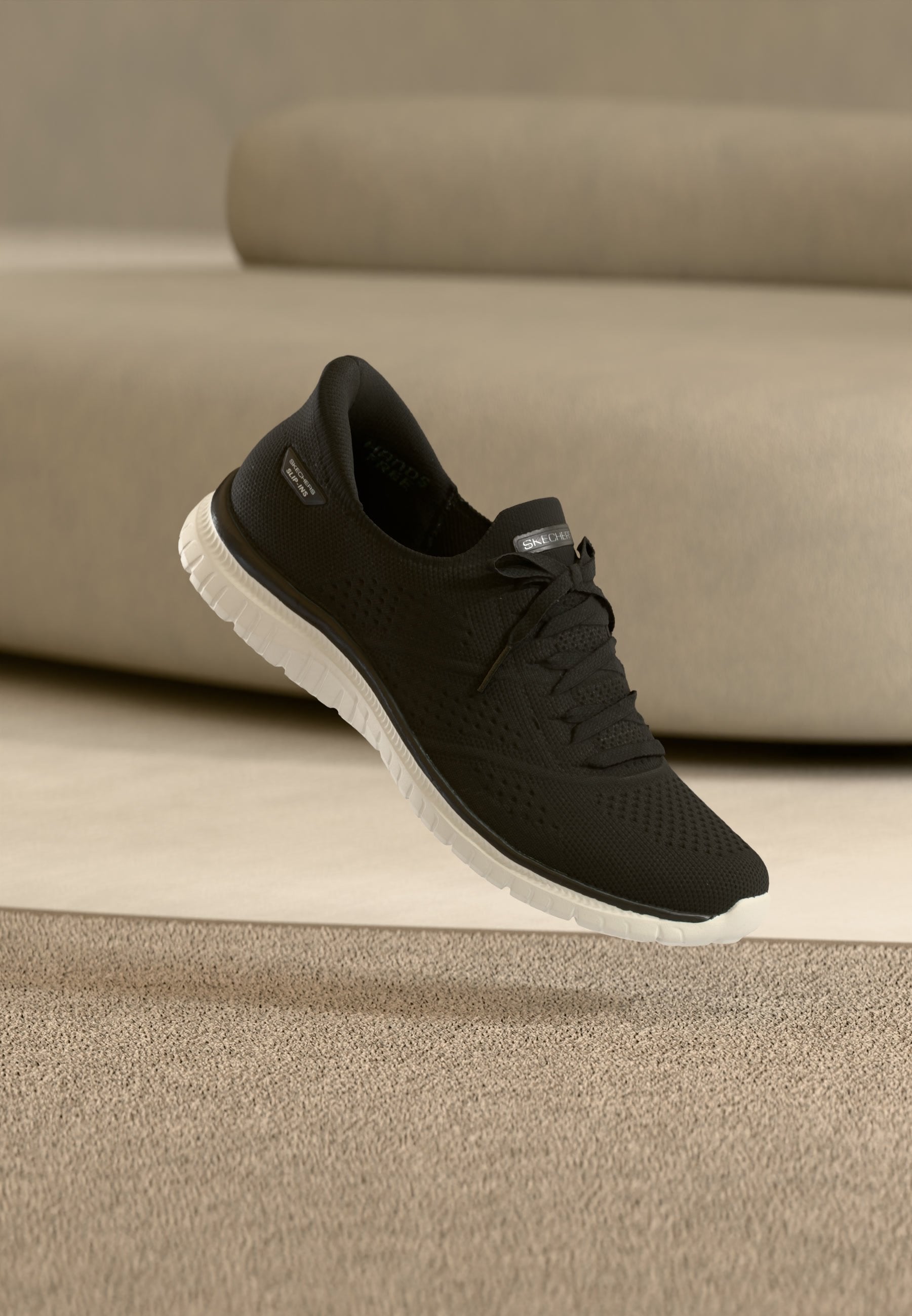 Schwarzer Strick-Sneaker mit weißer Sohle, Schnürsenkeln und Skechers-Logo auf der Zunge, schwebt über einem beigen Teppich vor einem beigen Sofa.