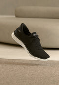 Schwarzer Strick-Sneaker mit weißer Sohle, Schnürsenkeln und Skechers-Logo auf der Zunge, schwebt über einem beigen Teppich vor einem beigen Sofa.