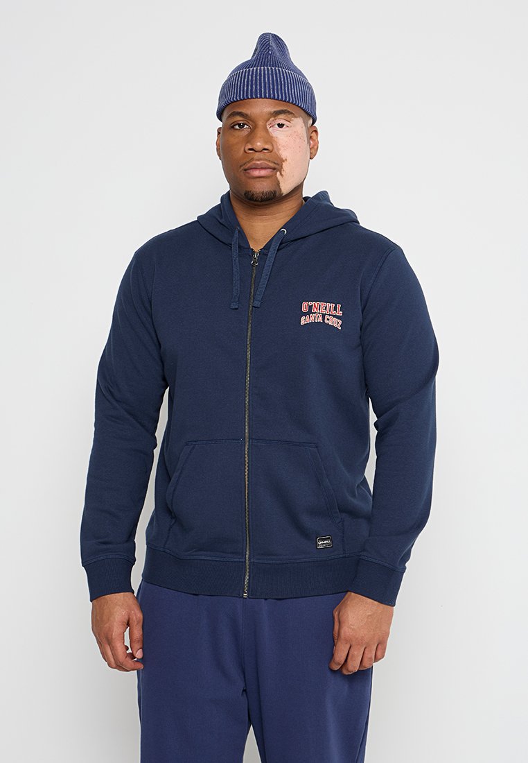 Marineblaue Zip-Hoodie mit vorderen Taschen, gerippten Bündchen und rotem O'Neill-Logo. Getragen mit blauen Sweatpants und einer blauen Strickmütze.