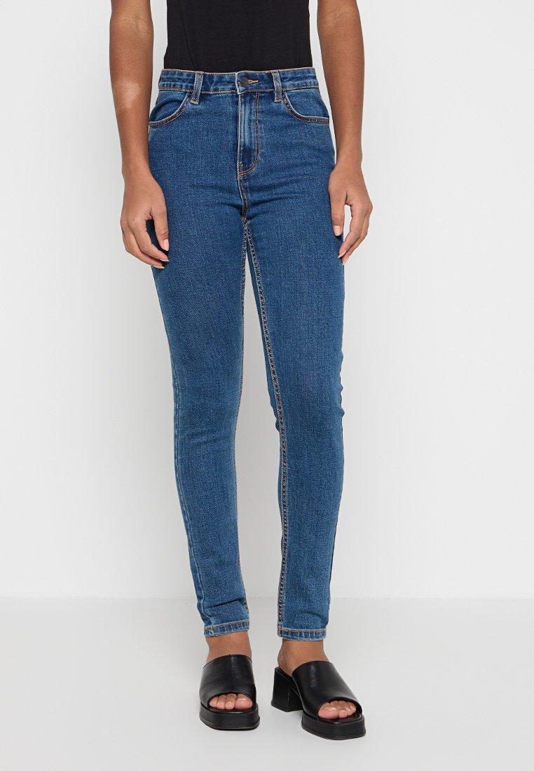 Frau trägt hochtaillierte blaue Skinny-Jeans, schwarzen ärmellosen Top und schwarze offene Sandaletten mit Absatz, steht vor einem einfarbigen Hintergrund.