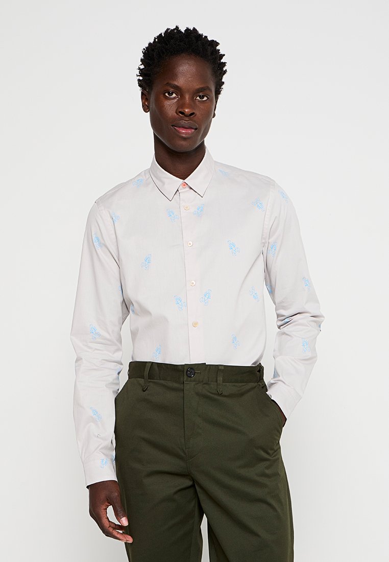 Lichtgraues Langarmhemd mit dezentem blauen Schmetterlingsmuster, Button-Down-Kragen und durchgehender Knopfleiste. Getragen mit dunkelgrünen Hosen.