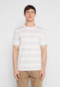 Mann steht, trägt weißes T-Shirt mit dünnen horizontalen grauen Streifen und hellbraune Hose vor schlichtem grauem Hintergrund.
