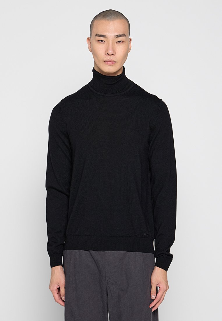 Schwarzer Rollkragenpullover mit gerippter Textur und langen Ärmeln. Enganliegendes Design mit flacher Strickkante. Getragen mit grauen Hosen.
