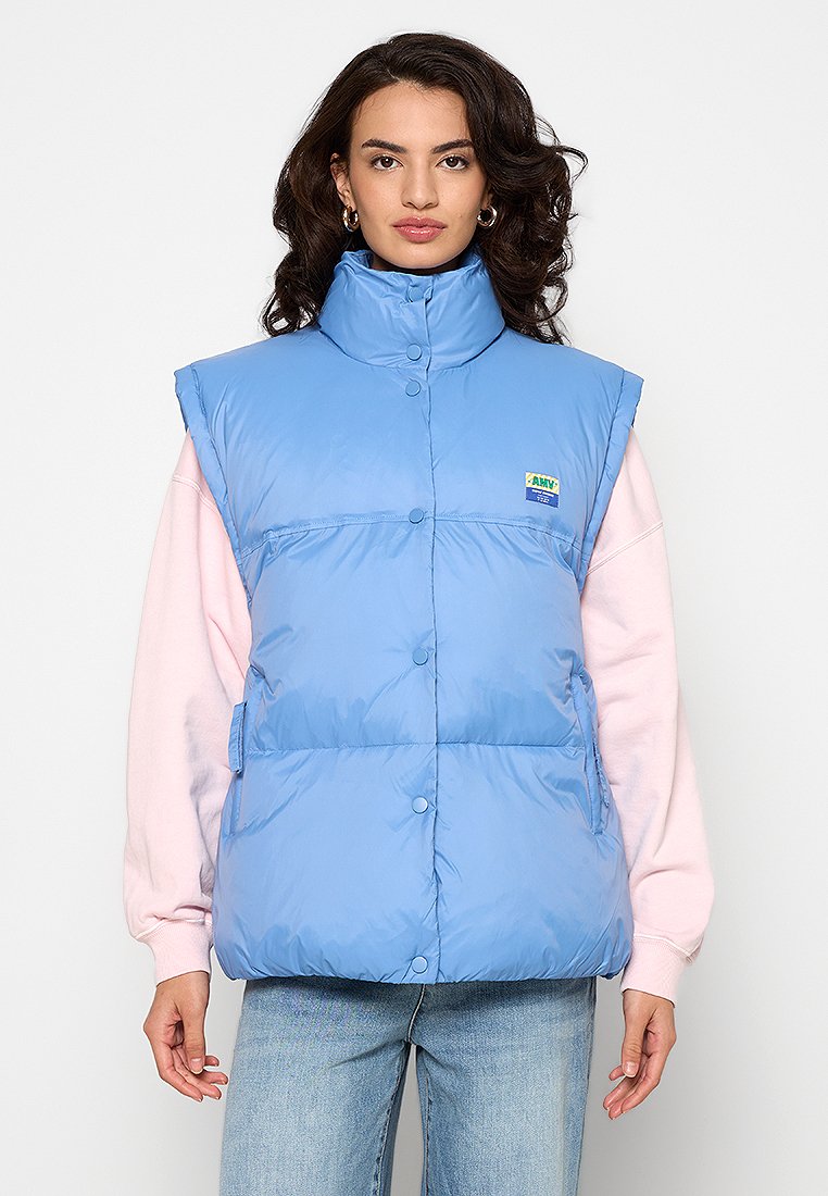 Hellblaues Puffer-Westen mit hohem Kragen, Druckknöpfen und seitlichen Taschen. Wird über einem weichen rosa Sweatshirt getragen, das ein Logopatch aufweist.