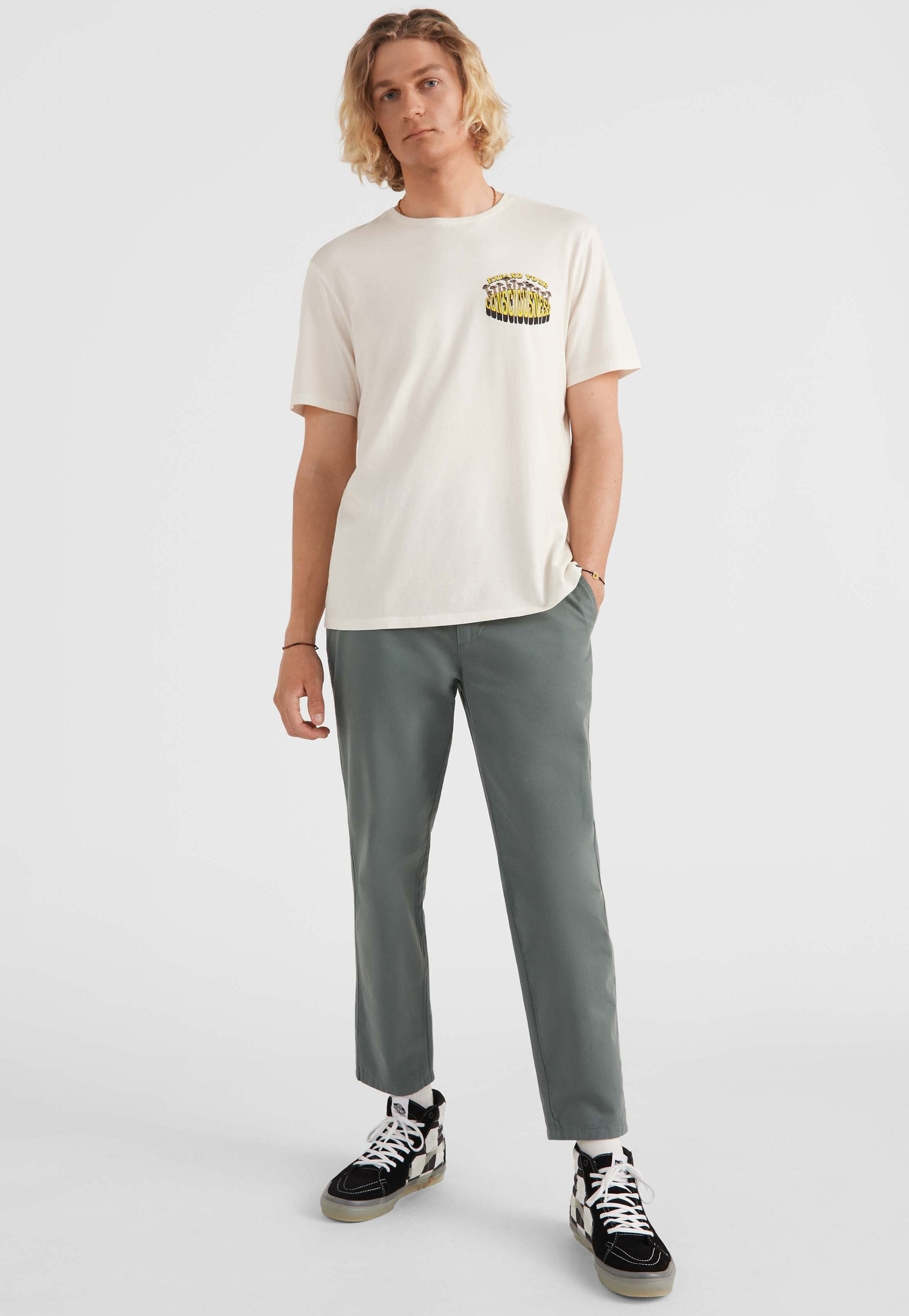 Cremefarbener Baumwoll-T-Shirt mit Grafikdruck auf der Brust, kombiniert mit locker sitzenden grünen Hosen und schwarz-weißen High-Top-Sneakern.