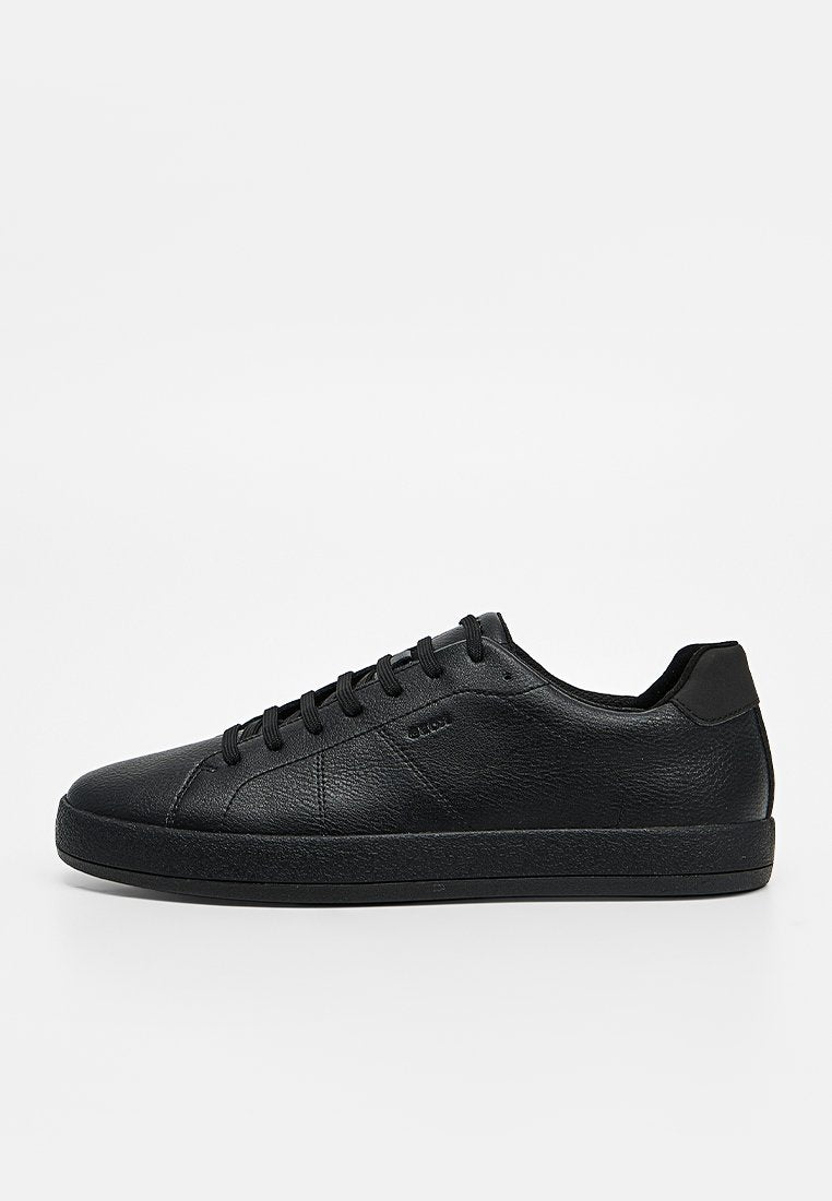 Schwarzer flacher Ledersneaker mit schwarzen Schnürsenkeln und profilierter Sohle, seitlich auf einem einfarbigen hellen Hintergrund dargestellt.