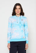 Frau mit schulterlangem braunem Haar trägt einen hellblauen Batik-Hoodie mit Kängurutasche und dunkler Hose und steht vor einem schlichten Hintergrund.