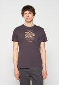 Junger Mann mit kurzen braunen Haaren, trägt ein dunkelgraues Schott-T-Shirt mit motorradbezogenem Text und lässige graue Hose, steht vor weißem Hintergrund.