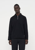 Mann mit dunkler Haut und geflochtenem Haar, der ein schwarzes Langarmshirt mit Reißverschlusskragen und schwarze Hose vor einfachem weißem Hintergrund trägt.