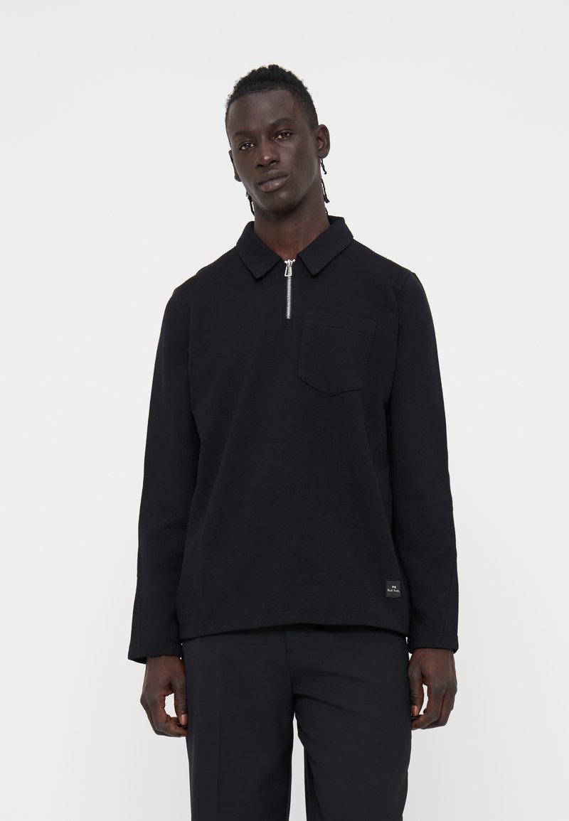Mann mit dunkler Haut und geflochtenem Haar, der ein schwarzes Langarmshirt mit Reißverschlusskragen und schwarze Hose vor einfachem weißem Hintergrund trägt.