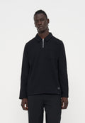 Mann mit dunkler Haut und geflochtenem Haar, der ein schwarzes Langarmshirt mit Reißverschlusskragen und schwarze Hose vor einfachem weißem Hintergrund trägt.