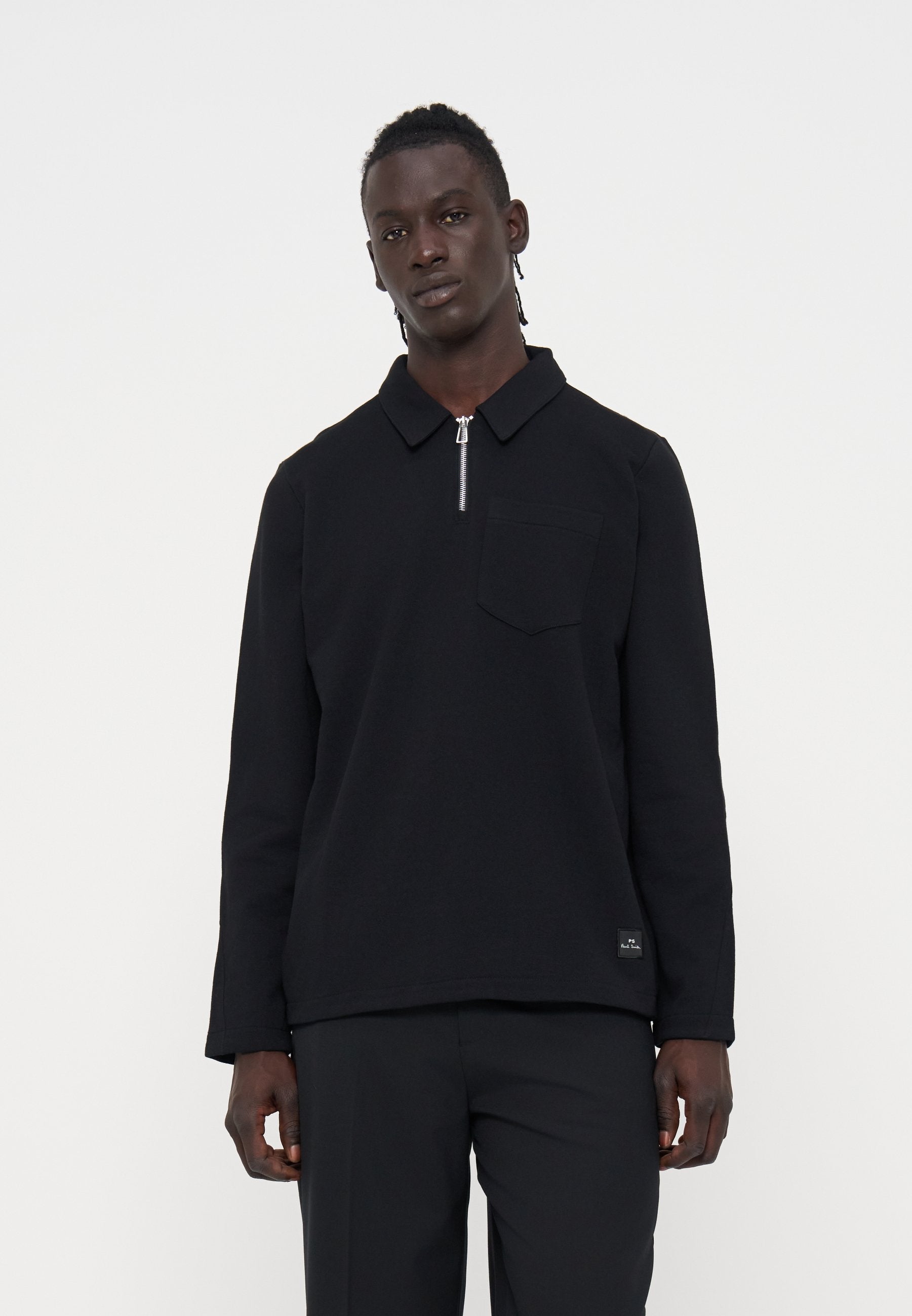 Mann mit dunkler Haut und geflochtenem Haar, der ein schwarzes Langarmshirt mit Reißverschlusskragen und schwarze Hose vor einfachem weißem Hintergrund trägt.