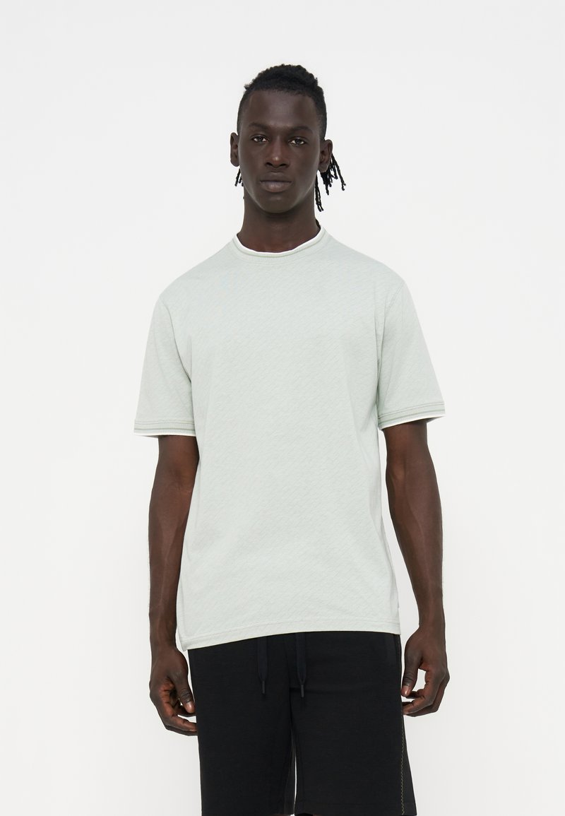 Mann mit kurzen Dreadlocks trägt ein schlichtes hellgraues Kurzarmshirt und lockere schwarze Hosen vor weißem Hintergrund.