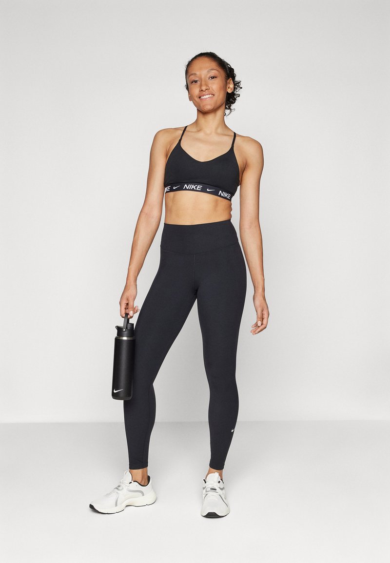 Schwarzer Sport-BH mit Nike-Logo, passende schwarze hoch taillierte Leggings und weiße Sportschuhe. Hält eine schwarze Wasserflasche.