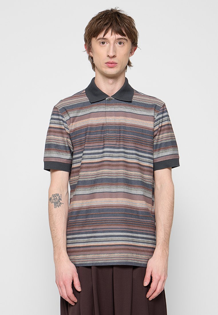 Junger Mann mit kurzen braunen Haaren, der ein kurzärmeliges gestreiftes Poloshirt in Brauntönen, Grau und Blau trägt und vor einem einfarbigen Hintergrund steht.