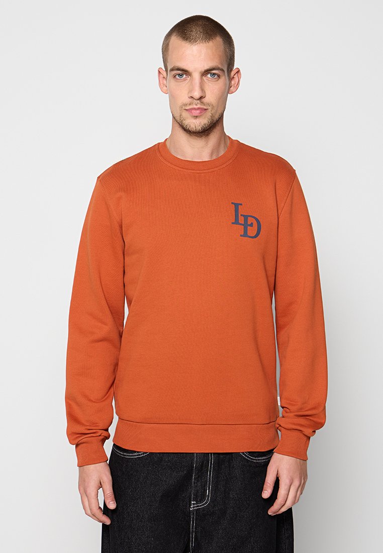 Rost-orange Langarm-Sweatshirt aus strukturiertem Stoff. Verfügt über einen Rundhalsausschnitt und ein marineblaues Logo auf der linken Brust. Mit gerippten Ärmelbündchen und Saum.