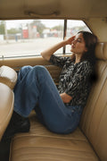 Schwarz-weiß gemusterte Bluse, blaue Jeans, sitzt in einem hellbraunen Lederautoinnenraum, mit Ausblick durch das Fenster und entspannter Haltung.