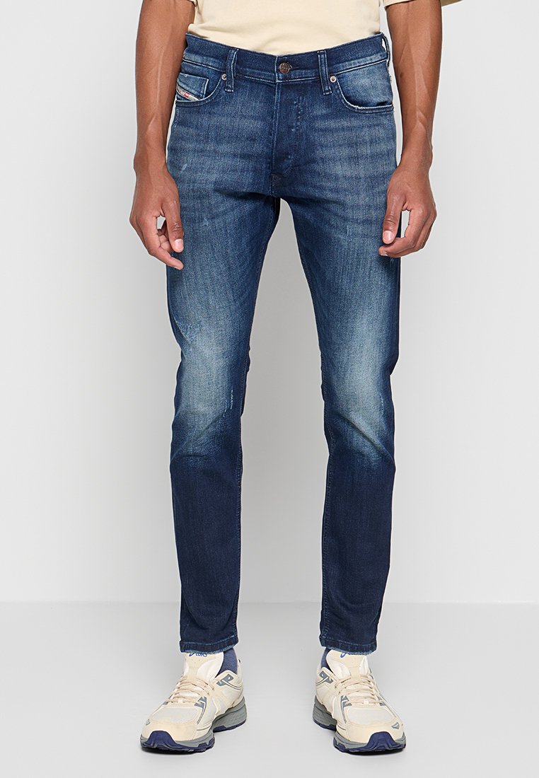 Dunkelblaue Jeans im Slim-Fit-Schnitt mit leichten Abnutzungen an den Oberschenkeln, fünf Taschen und einem Knopfverschluss. Getragen mit hellen Sneakers.