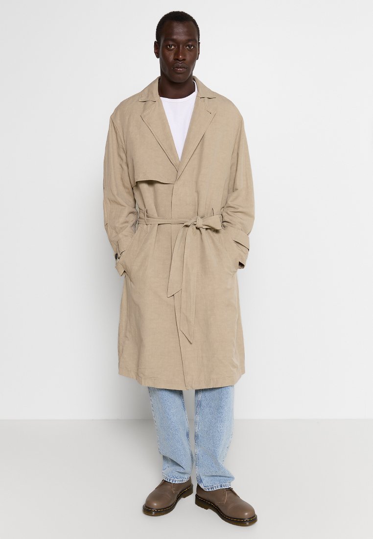 Beige Leinen-Trenchcoat mit Gürtel, langen Ärmeln und Fronttaschen, kombiniert mit hellblauen Jeans und braunen Schuhen. Minimalistisches Design.