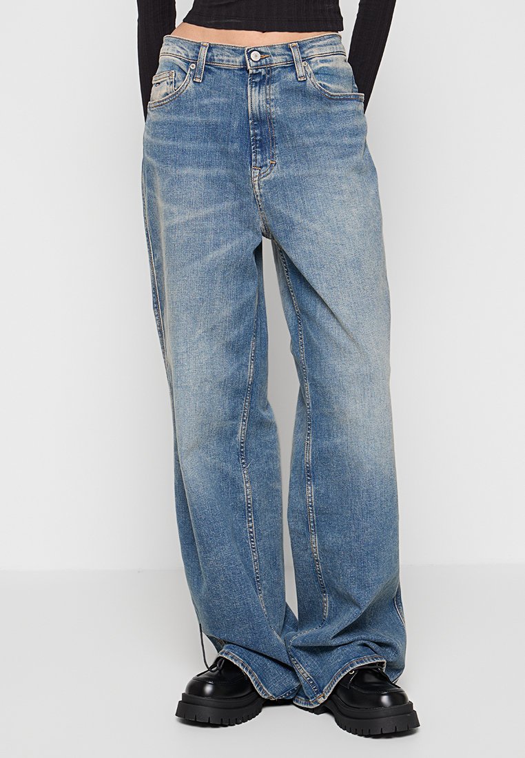 Person, die lockere, ausgebleichte blaue Jeans mit hoher Taille und schwarze Schuhe mit dicker Sohle trägt, steht vor einem schlichten hellen Hintergrund.