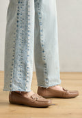 Braune Lederloafers mit strukturiertem Finish und kleinem metallic Akzent, kombiniert mit hellblauen, ausgefransten Jeans, auf einem Holzboden.