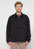 Mann mit lockigem Haar, der einen schwarzen gesteppte Pullover mit einem Viertel-Reißverschluss und beigen Hosen trägt, steht vor einem schlichten Hintergrund.