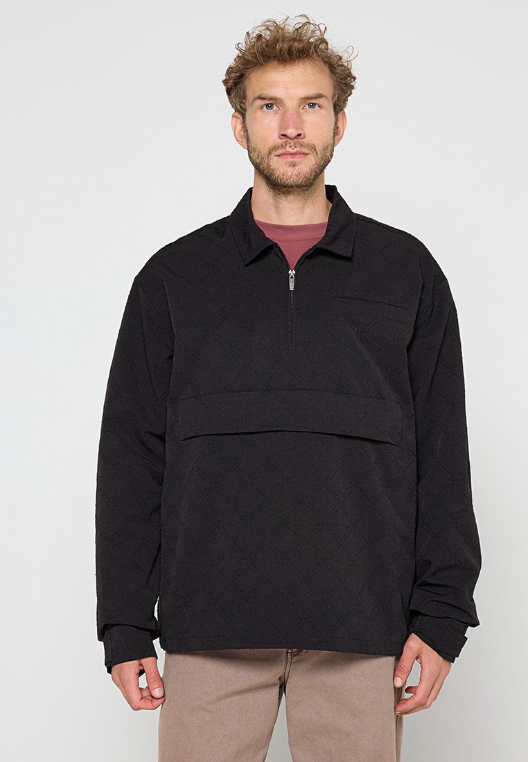Mann mit lockigem Haar, der einen schwarzen gesteppte Pullover mit einem Viertel-Reißverschluss und beigen Hosen trägt, steht vor einem schlichten Hintergrund.