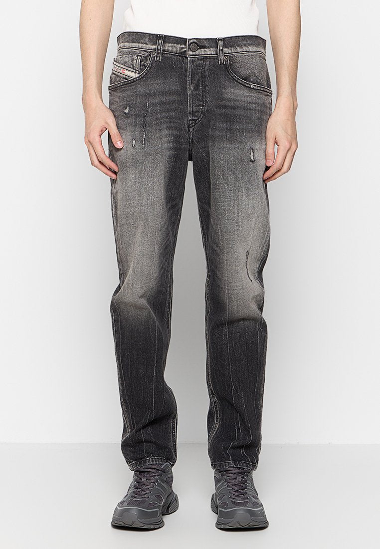 Schwarze Denim-Jeans mit einem distressed Finish, verblasster Textur, zwei vorderen Taschen und einem Knopfverschluss. Kombiniert mit grauen Sportschuhen.