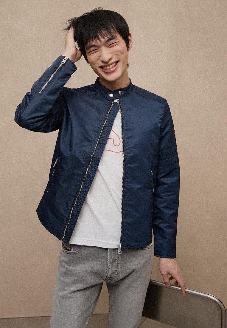 Marineblaue Jacke mit Reißverschluss vorne und hohem Kragen, mit glatter, glänzender Oberfläche und Seitentaschen. Wird über einem weißen T-Shirt getragen.
