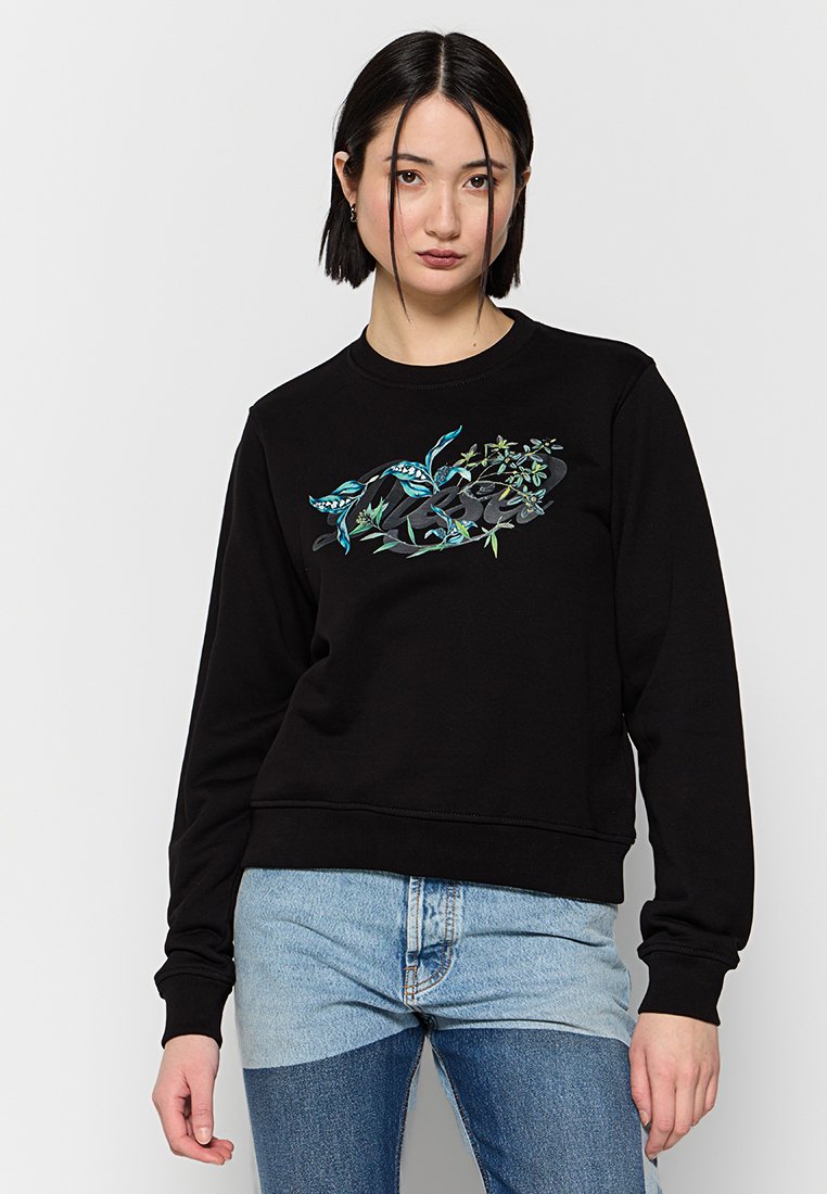 Schwarzes Sweatshirt mit einem Blumenmuster in Blau und Grün auf der Brust, Rundhalsausschnitt und gerippten Bündchen. Kombiniert mit hellen Denim-Hosen.