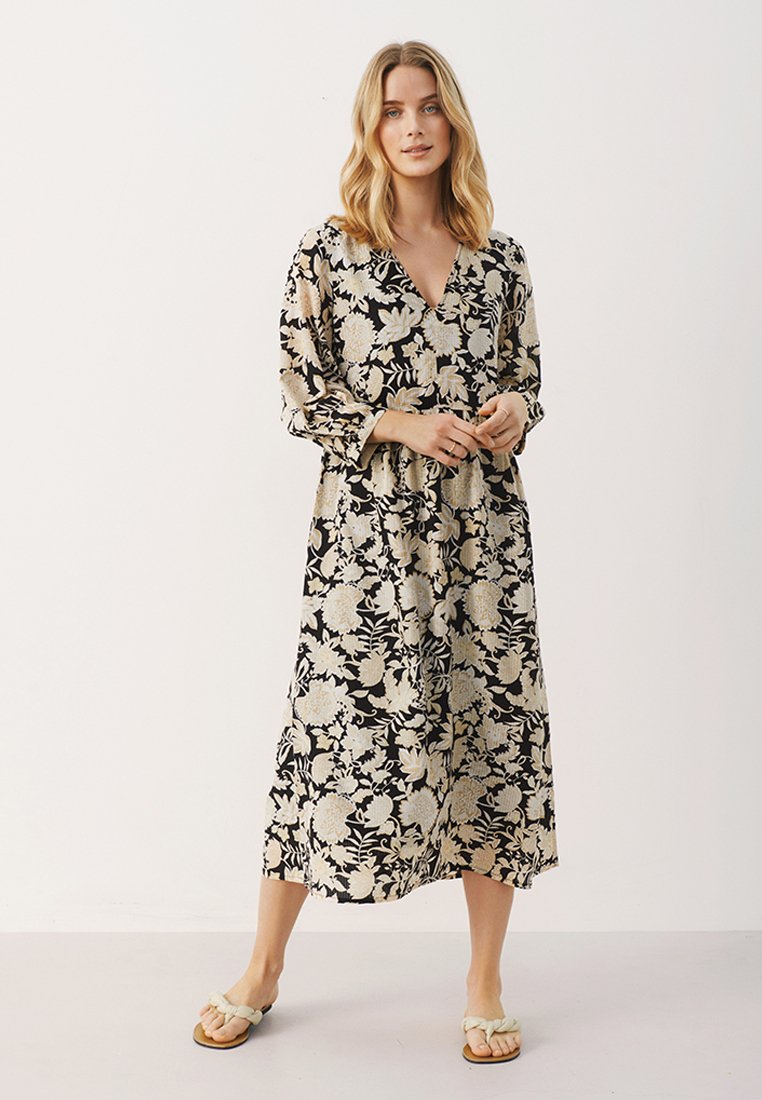 Schwarzes Kleid mit cremefarbenem Blumenmuster, V-Ausschnitt, langen Ärmeln und lockerem Schnitt, das bis zur Mitte der Wade reicht. Kombiniert mit hellen Sandalen.