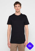 Junger Mann trägt ein schlichtes schwarzes kurzärmeliges T-Shirt und beige Hosen, steht vor einem einfarbigen hellgrauen Hintergrund.