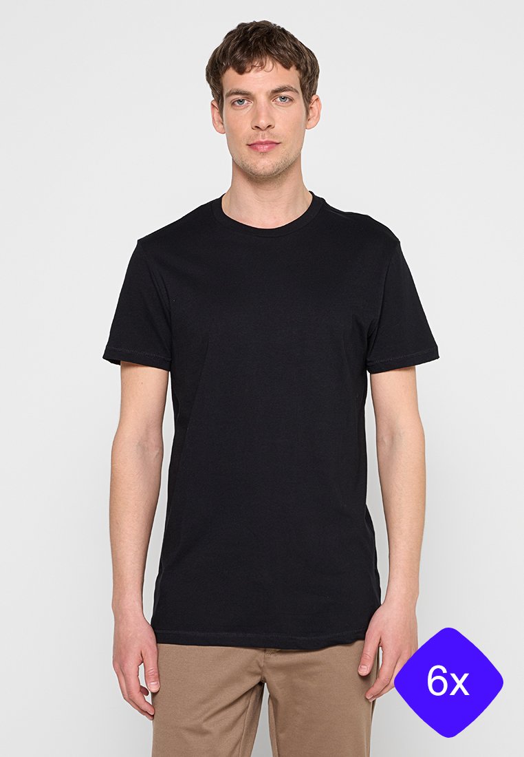 Junger Mann trägt ein schlichtes schwarzes kurzärmeliges T-Shirt und beige Hosen, steht vor einem einfarbigen hellgrauen Hintergrund.