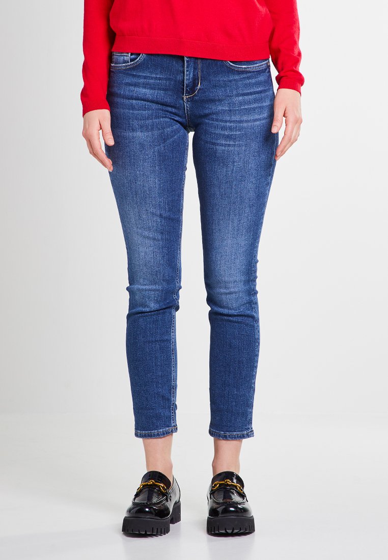 Dünne blaue Denim-Jeans mit einem mittelhohen Bund, Fünf-Taschen-Design und subtiler, verblasster Textur. Kombiniert mit schwarzen Schuhen mit dicker Sohle.