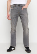Mann in ausgeblichenen grauen Jeans und schwarzen Sneakers mit weißen Streifen, der mit einer Hand in der Tasche vor weißem Hintergrund steht.
