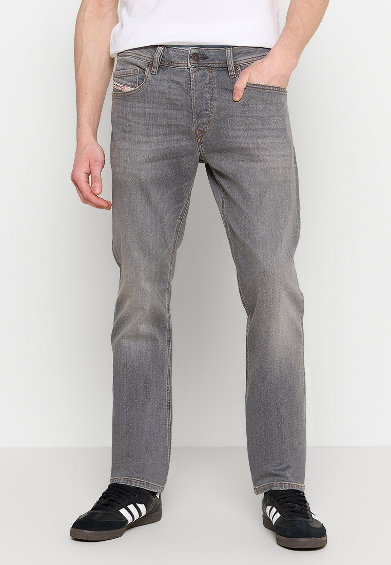 Mann in ausgeblichenen grauen Jeans und schwarzen Sneakers mit weißen Streifen, der mit einer Hand in der Tasche vor weißem Hintergrund steht.