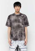 Übergroßes graues T-Shirt mit einem verbrannten Finish, das ausgefranste Kanten und ein kleines Logoschild aufweist. Getragen mit weißen karierten Cargo-Hosen.