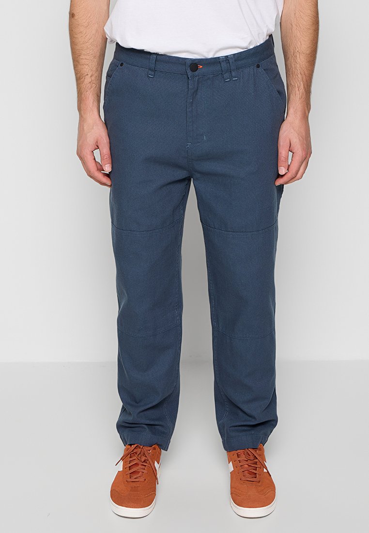 Mann trägt dunkelblaue gerade geschnittene Hose mit Seitentaschen und verbrannte orangefarbene Sneakers, steht vor einem weißen Hintergrund.