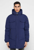 Mann in einer dunkelblauen gepolsterten Winterjacke mit Kapuze, zwei Klappentaschen auf der Brust und seitlichen Reißverschlusstaschen, der vor einem schlichten Hintergrund steht.