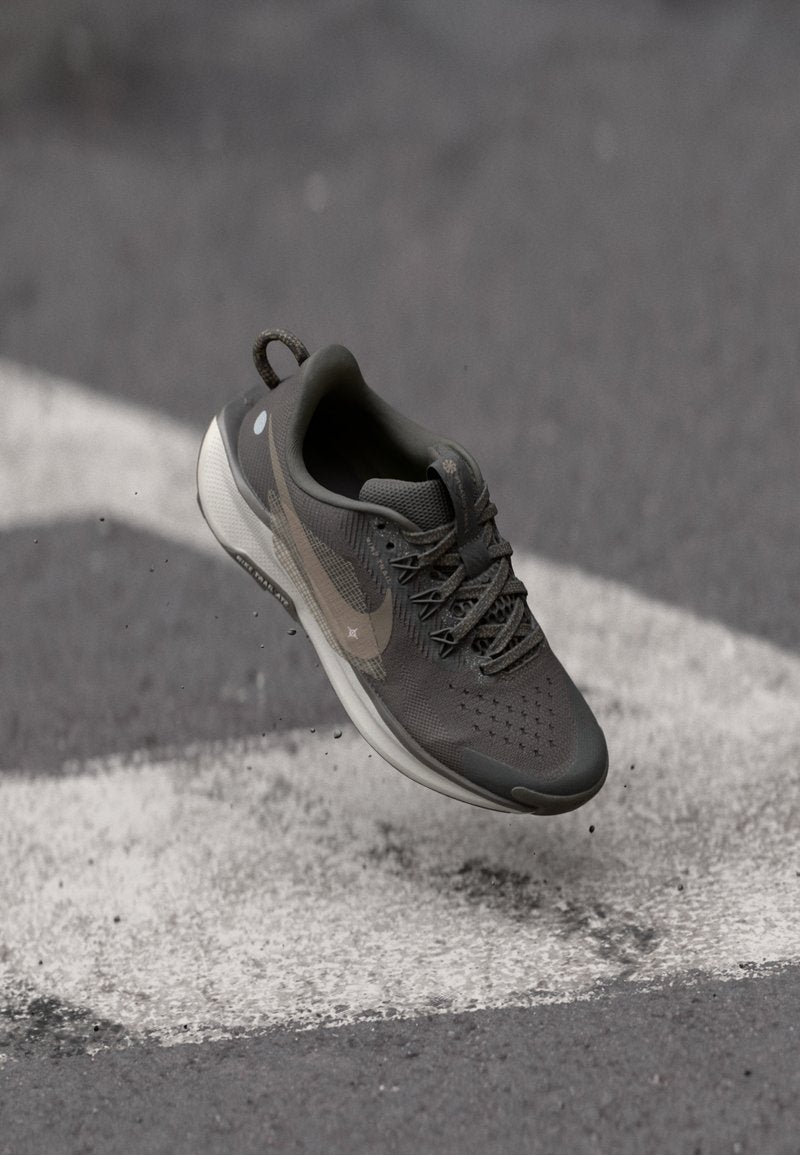 Graue Sportschuhe mit einer strukturierten Oberseite, Schnürverschluss und einer weißen Sohle. Verfügt über ein dezentes Logo und reflektierende Akzente. Staubpartikel sichtbar.