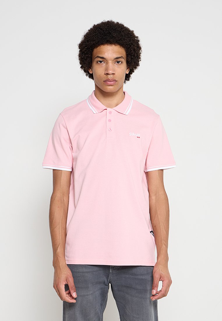 Junger Mann mit lockigem Haar, der ein hellrosa Polohemd und graue Hose trägt, steht vor einem einfarbigen grauen Hintergrund.